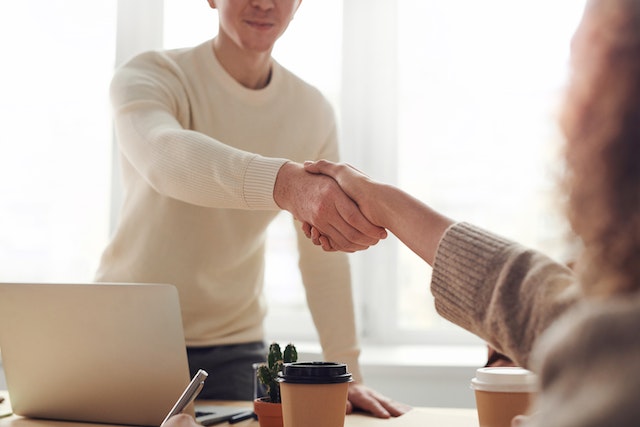two people shaking hands over a laptop and coffee in an office